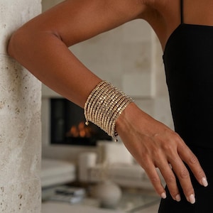 Può includere: Un bracciale a fascia larga composto da molteplici file di piccole perline metalliche quadrate color oro. Il bracciale presenta un design flessibile a più fili che avvolge il polso, creando un aspetto strutturato e uniforme con una finitura lucida.