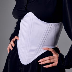 May include: A white corset with vertical stitching details. The corset is worn over a black top and dress. The corset has a curved, pointed bottom edge and a smooth, fitted design.