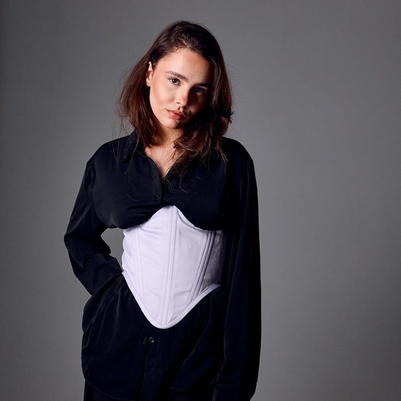 May include: A white corset with vertical seams is worn over a black button-down shirt. The corset has a curved neckline and a fitted silhouette. The model is facing the camera with a neutral expression. The background is a solid gray.