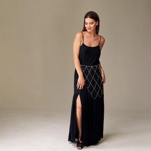 May include: A black maxi dress with spaghetti straps and a thigh-high slit. A silver chain belt with a diamond pattern is worn around the waist. The dress is made of a flowing fabric.