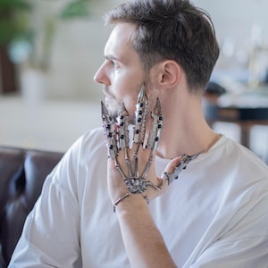 May include: A silver-colored articulated hand armor with sharp, claw-like extensions and black accents. The gothic-inspired accessory features a skeletal design, with each finger and knuckle detailed. The armor is worn on the hand, showcasing a unique, edgy style.