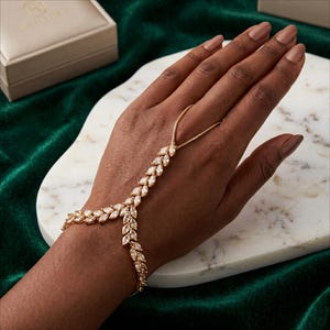 Puede incluir: Una pulsera de mano dorada con cristales transparentes tallados en forma de marquesa. La pulsera presenta una delicada cadena que se extiende hasta un anillo para el dedo. La joya se exhibe en una mano sobre un fondo de terciopelo verde.