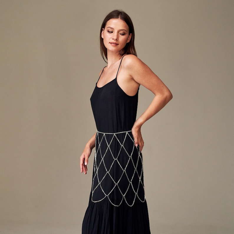 May include: A black dress with spaghetti straps and a tiered skirt. A silver-colored body chain with a diamond pattern is worn around the waist. The dress is a solid color and appears to be made of a soft fabric.