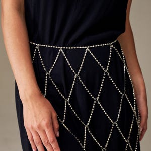 May include: A silver body chain with a diamond pattern. The chain is made of small, sparkling rhinestones and is worn over a black dress. The chain is designed to be worn around the waist and hips, adding a touch of glamour.