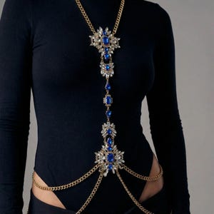 Puede incluir: Una cadena de cuerpo dorada con un colgante central con grandes piedras preciosas azules y cristales transparentes más pequeños. La cadena se extiende por el torso y se conecta a una cadena de cintura. La joya se usa sobre un body negro.