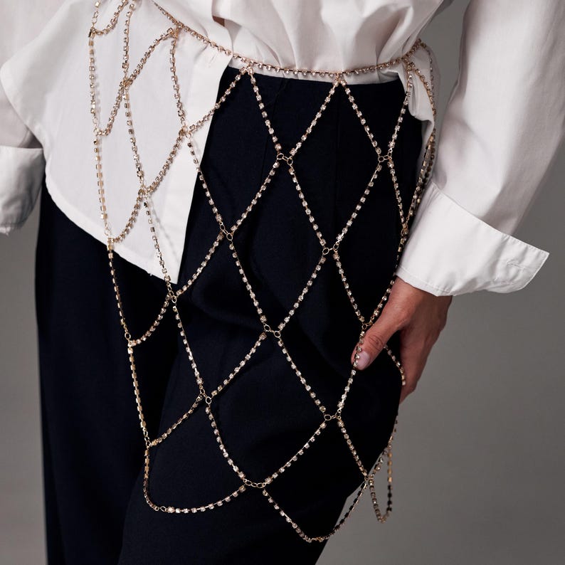 May include: Gold-toned body chain with a diamond pattern, draped over black pants. The chain is made of small, sparkling rhinestones and is worn over a white button-down shirt. The chain is designed to be worn around the waist and over the leg.