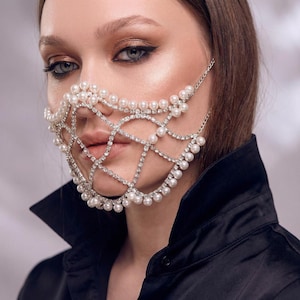 Può includere: Una maschera per il viso argentata dal design delicato, con perle finte e cristalli scintillanti. La maschera è collegata da catene che si agganciano alle orecchie. La modella indossa una camicia di raso nera.