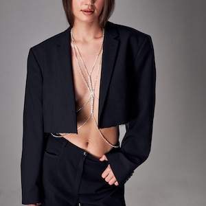 Peut inclure: Un blazer noir court avec un ourlet brut, associé à un pantalon noir. Une chaîne de corps argentée à plusieurs brins orne le torse. L'ensemble est posé sur un fond gris neutre.