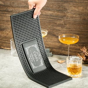 Puede incluir: Una alfombrilla de bar de goma negra con un letrero rectangular personalizado que dice "Welcome Caleb's Bar - Enjoy the day!". La alfombrilla es curva y tiene una superficie texturizada. La imagen también muestra un cóctel y un vaso de whisky.