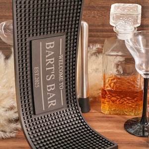 May include: A personalized bar setup featuring a black rubber bar mat with the text "BART'S BAR EST.2025". Includes a glass decanter, cocktail shaker, martini glass, and a jigger. The scene is set on a wooden surface.
