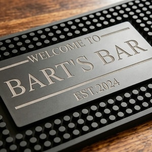 Puede incluir: Una alfombrilla de bar negra con una placa rectangular grabada que dice "WELCOME TO BART'S BAR EST. 2024". La alfombrilla tiene una superficie texturizada con pequeñas protuberancias redondeadas.