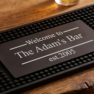 May include: A black bar mat with a rectangular metal sign that reads "Welcome to The Adam's Bar est.2005" in white lettering. The mat has a textured surface and is on a wooden bar.