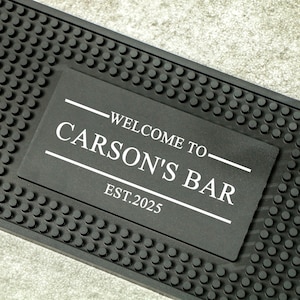 Può includere: Tappetino da bar nero con un cartello rettangolare con la scritta bianca "WELCOME TO CARSON'S BAR EST.2025". Il tappetino ha una superficie strutturata con piccoli rilievi circolari. Il cartello è centrato.