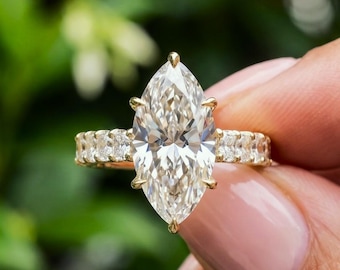 May include: A gold ring featuring a large marquise-cut center stone held by six prongs, with a band encrusted with smaller round-cut stones along the sides. The metal band has a polished finish and the stones exhibit a clear, reflective quality.