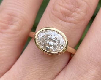 May include: A gold ring featuring a single oval-cut clear gemstone set in a smooth bezel setting. The band is thin and polished, showcasing a minimalist design that highlights the central stone's facets and clarity in a classic yellow gold finish.