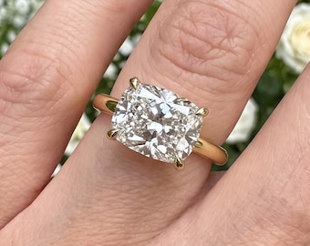 May include: A gold ring featuring a large, rectangular cushion-cut clear gemstone set with four prongs. The band is a simple, polished gold metal. The stone has multiple facets that reflect light, creating a bright, sparkling appearance on the finger.