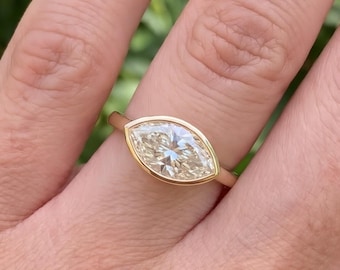 May include: A gold ring featuring a marquise-cut diamond set in a smooth bezel setting. The diamond has a light champagne hue and is positioned horizontally across the finger. The band is a simple, polished gold metal with a rounded profile.