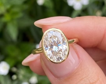 May include: A gold ring featuring a large, oval-cut, clear faceted gemstone set in a smooth, polished gold bezel setting. The band is thin and made of solid gold, showcasing a clean, reflective surface that catches the light.