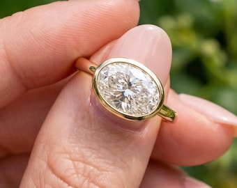 May include: A gold ring featuring a large, oval-cut, clear gemstone set in a smooth, polished bezel setting. The band is thin and made of yellow gold, providing a clean, continuous frame around the faceted stone that reflects light.