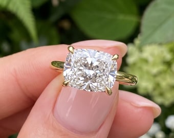 May include: A gold ring featuring a large, cushion-cut, clear faceted gemstone held by four prongs. The band is a simple, polished gold metal. The ring is positioned against a backdrop of green foliage and small white flowers.