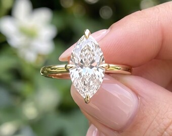 May include: A marquise-cut diamond ring set on a polished yellow gold band. The center stone is held in place by a four-prong setting, showcasing a clear, faceted gemstone that reflects light with a brilliant, elongated shape.