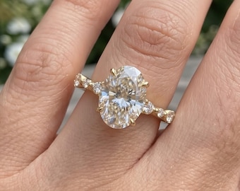 May include: A gold ring featuring a large oval-cut center stone held by four prongs, flanked by smaller round-cut stones set along the band. The metal band has a polished finish, and the design incorporates multiple accent stones on each side.