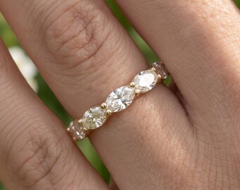 May include: A gold band ring featuring a series of five oval-cut clear stones arranged horizontally along the top. The stones are set in a shared prong setting, creating a continuous line of sparkle across the finger with a polished metal finish.