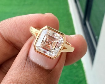 May include: A gold ring featuring a large, square-cut, clear gemstone set in a bezel setting. The band is smooth and polished, showcasing a clean, geometric design with a prominent center stone that reflects light across its faceted surface.