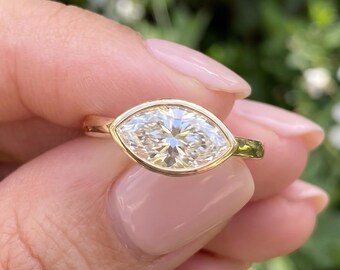 May include: A marquise-cut gemstone ring set in a polished yellow gold bezel setting. The elongated, pointed oval stone features multiple facets that reflect light, secured by a smooth, continuous gold band that encircles the finger.