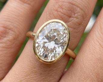 May include: A gold ring featuring a large, oval-cut, clear faceted gemstone set in a smooth, polished bezel setting. The band is thin and circular, crafted from a warm-toned yellow gold metal that encircles the central oval stone securely.