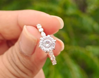 May include: A silver-toned ring featuring a large, round, faceted clear stone as the centerpiece, flanked by smaller, round and pear-shaped clear stones set along the band, creating a continuous sparkling effect across the top of the ring.
