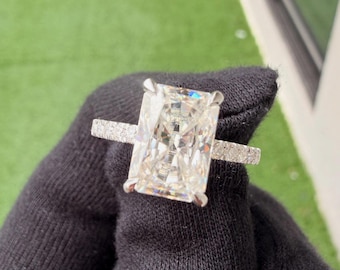 May include: A radiant-cut solitaire ring featuring a large, rectangular, clear gemstone held by four silver-toned prongs. The thin band is encrusted with small, round, sparkling stones along the visible portion of the shank, creating a refined look.