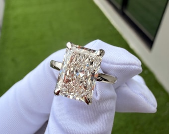 May include: A radiant-cut diamond ring featuring a large, rectangular center stone with brilliant facets, held by four silver-toned metal prongs on a polished, smooth, thin silver-toned metal band, showcasing high clarity and light reflection.
