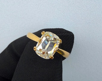 May include: A gold ring featuring a large, rectangular cushion-cut clear gemstone set with four prongs. The band is smooth and polished gold, showcasing a classic design that highlights the faceted stone and its reflective, brilliant surface.