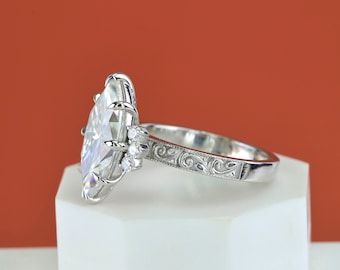 May include: A silver-toned ring featuring a large marquise-cut center stone held by multiple prongs. The band is decorated with intricate scrollwork engravings and includes three small round accent stones on each side of the main setting.