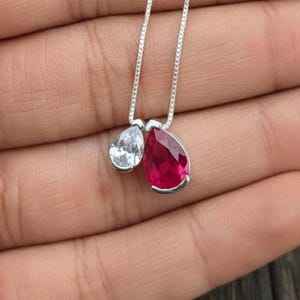 Könnte beinhalten: Eine silberne Halskette mit zwei tropfenförmigen Anhängern. Ein Anhänger ist ein klarer, weißer Edelstein, der andere ein leuchtend roter Edelstein. Die Anhänger hängen an zarten Silberketten.