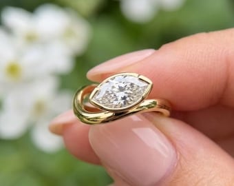 May include: A gold ring featuring a marquise-cut diamond set in a bezel setting. The band has a smooth, polished finish and a bypass design that curves around the central stone, creating a continuous, flowing look in this piece of jewelry.