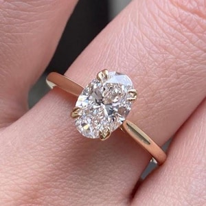 May include: A gold engagement ring with an oval-cut diamond. The diamond is held in place by four prongs on a slender, polished gold band. The ring is displayed on a finger.