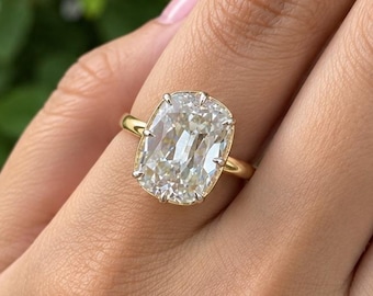 May include: A gold ring featuring a large, elongated cushion-cut clear gemstone set in a four-prong setting. The band is a simple, polished gold metal. The stone has a faceted surface that reflects light, creating a bright, sparkling appearance.