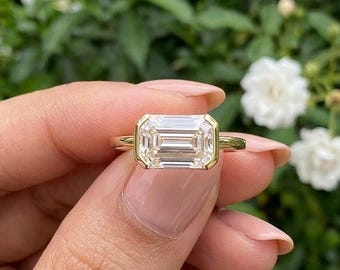 May include: A gold ring featuring a large, rectangular emerald-cut clear gemstone set in a smooth, polished gold bezel setting. The band is a simple, thin, polished gold wire that connects to the base of the rectangular stone setting.