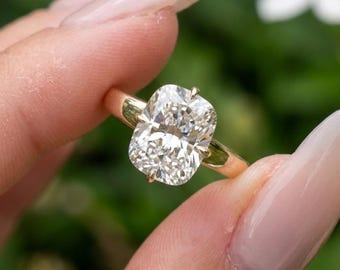 May include: A gold engagement ring featuring a large, rectangular-cut diamond. The diamond is held by four prongs and set on a delicate gold band. The ring is held up against a blurred green background.