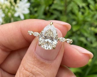 May include: A gold ring featuring a large pear-shaped center stone held by four prongs, flanked by three small marquise-cut stones on each side of the band. The metal band is thin and polished, showcasing a classic design with clear, faceted stones.