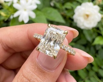 May include: A gold ring featuring a large, rectangular radiant-cut center stone held by four prongs. The band is crafted from yellow gold and is accented with a row of small, round-cut pavé diamonds extending along the sides of the ring shank.