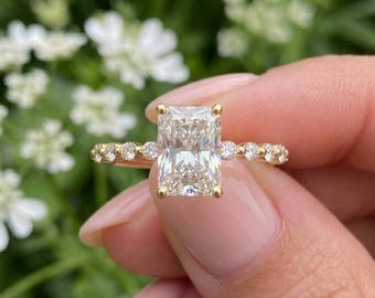 May include: A radiant-cut diamond engagement ring with a gold band. The ring features a large, rectangular diamond as the centerpiece, flanked by smaller, round diamonds along the band. The ring is held up against a blurred background of white flowers and green foliage.