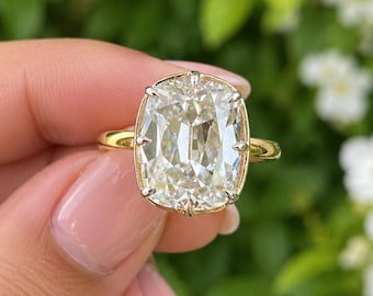 May include: A gold ring featuring a large, rectangular cushion-cut clear gemstone set in a prong setting. The stone has a faceted surface that reflects light, and the band is a simple, polished gold metal. The ring is held up against a natural backdrop.