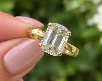 May include: A gold ring featuring a large, rectangular, emerald-cut clear gemstone held in place by four prongs. The band is made of polished yellow gold, providing a smooth and simple setting for the prominent, faceted center stone.