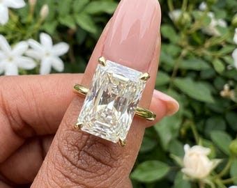 May include: A gold ring featuring a large, rectangular radiant-cut clear gemstone held by four prongs. The stone has a faceted surface that reflects light, and the band is a simple, thin gold wire that supports the prominent central setting.
