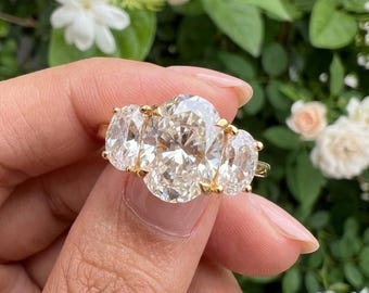 May include: A three-stone ring featuring a large central oval-cut diamond flanked by two smaller oval-cut diamonds, all set in a yellow gold band. The stones are clear and faceted, reflecting light with a brilliant sparkle against the metal setting.