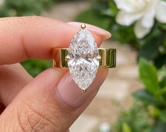 May include: A gold ring featuring a large, clear, marquise-cut gemstone set in a four-prong setting. The band is wide, smooth, and polished, showcasing a reflective metallic finish that contrasts with the faceted, brilliant-cut center stone.