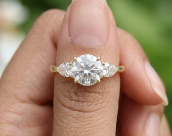 May include: A gold ring featuring a large, round, brilliant-cut center stone flanked by two smaller, pear-shaped side stones. The setting is crafted from polished yellow gold, showcasing a classic three-stone design with clear, faceted gemstones.